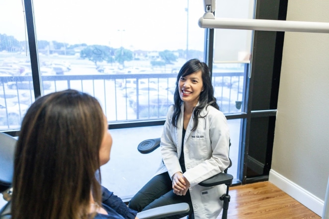 Two women having a conversation in a modern dental office with city views through windows.