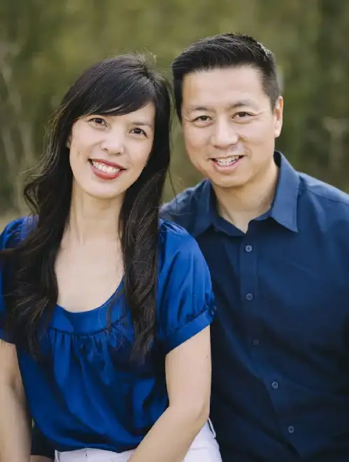 Two smiling people wearing blue shirts pose together outdoors with green foliage background.
