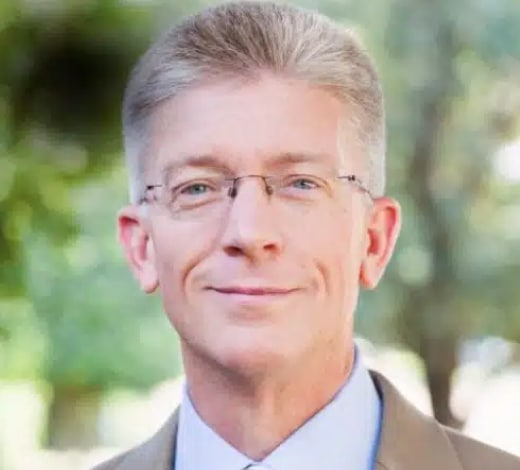 Professional headshot of middle-aged man wearing glasses and business attire against blurred outdoor background.
