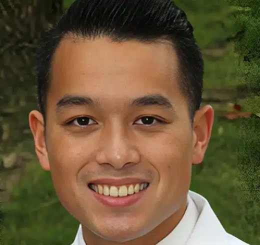 Professional headshot of smiling male dentist against green foliage background for dental practice.