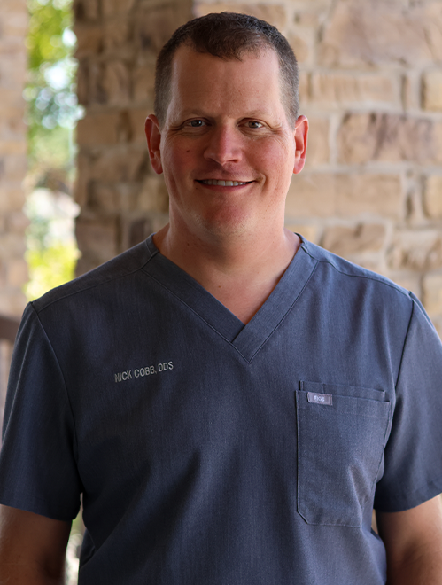 Male dentist in blue scrubs smiling at camera against brick wall background.