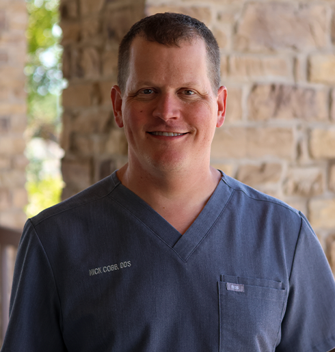 Male dentist in navy scrubs smiling at camera against brick wall background.