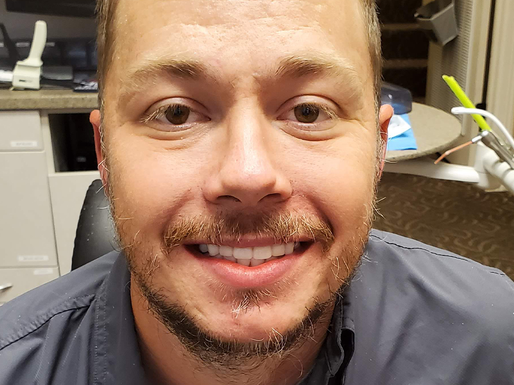 Smiling man in dental office wearing blue scrubs, indoor clinic setting with cabinets visible behind.