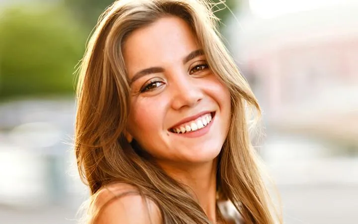 Smiling woman with long brown hair in outdoor setting, representing Denton Smiles Dentistry patient testimonial.