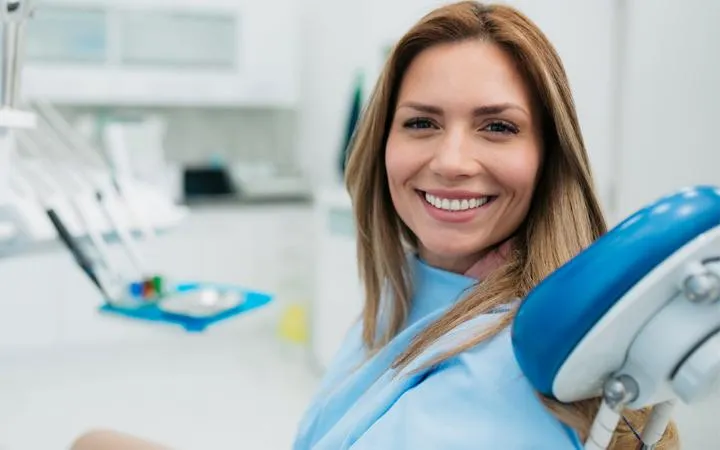 Smiling female dental professional in blue uniform at modern dental office with equipment visible.