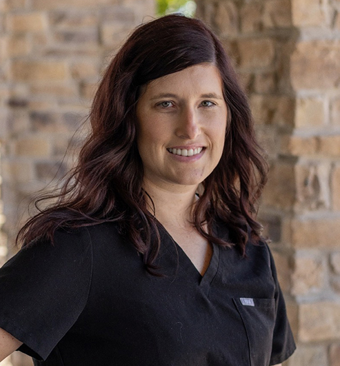 Woman in black scrubs smiling at camera against brick wall background.
