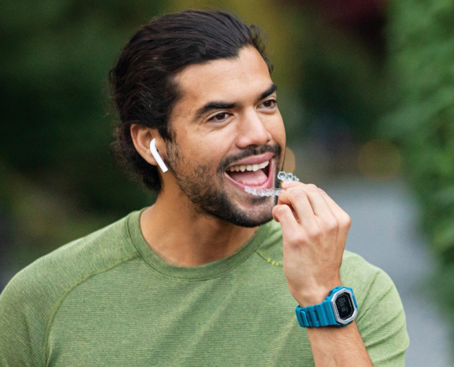 Man in green shirt smiling while brushing teeth outdoors, wearing blue smartwatch.