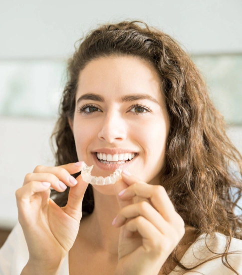 Woman holding clear dental aligners, smiling in bright clinical setting.