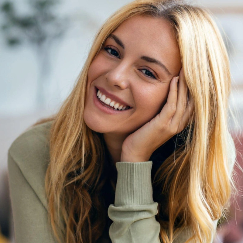 Smiling woman with blonde hair in casual green sweater, hand to face, bright indoor setting.