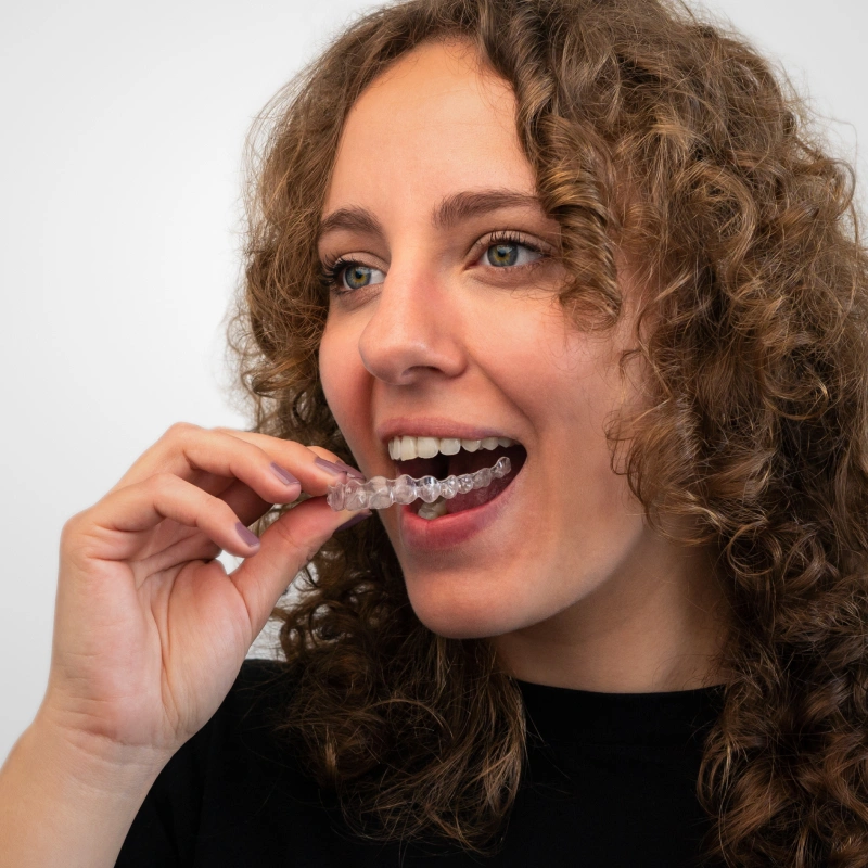 Woman with curly brown hair smiling while inserting clear dental aligners against white background.