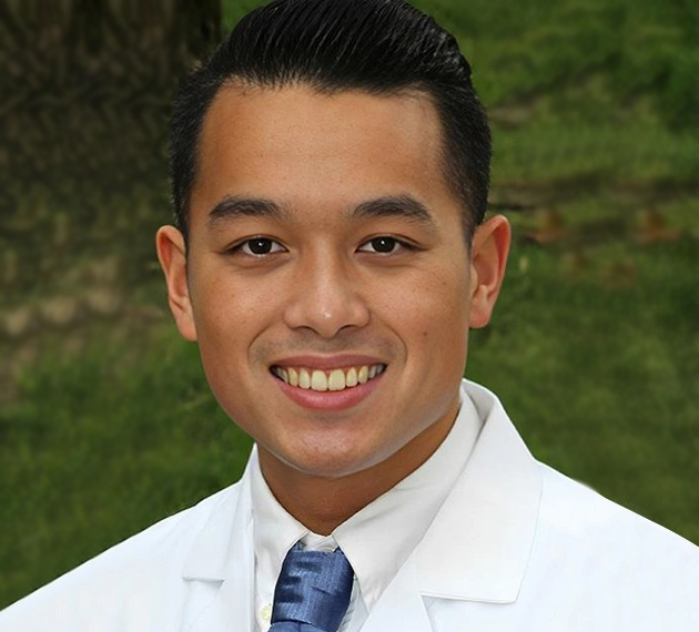 Professional headshot of male dentist in white coat and blue tie smiling at camera against green background.