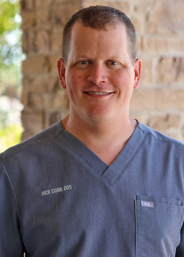 Male dentist wearing blue scrubs smiling at camera in dental office setting.