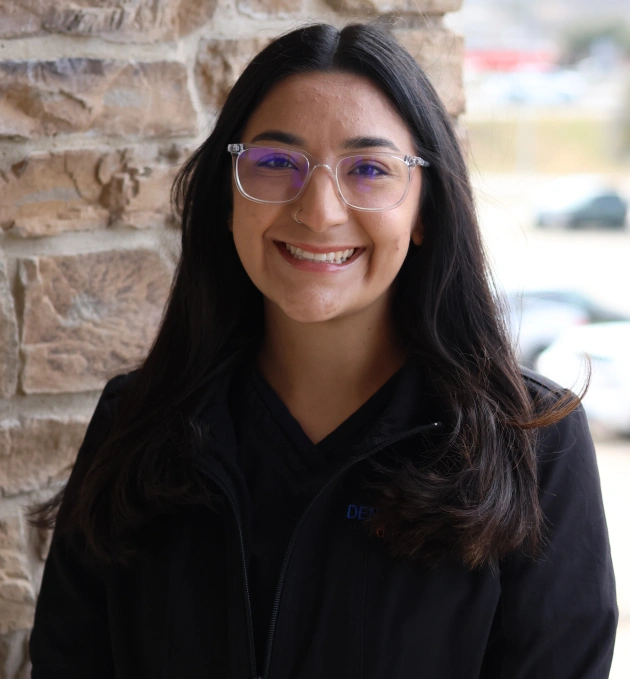 Young woman wearing purple glasses and black hoodie smiling at camera against brick wall.