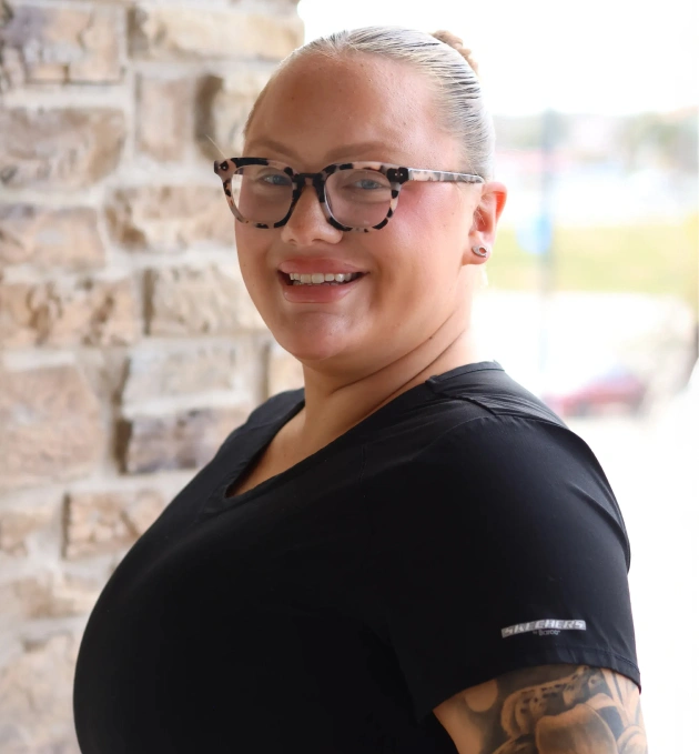 Professional dental staff member smiling at camera wearing glasses and black shirt against brick wall.