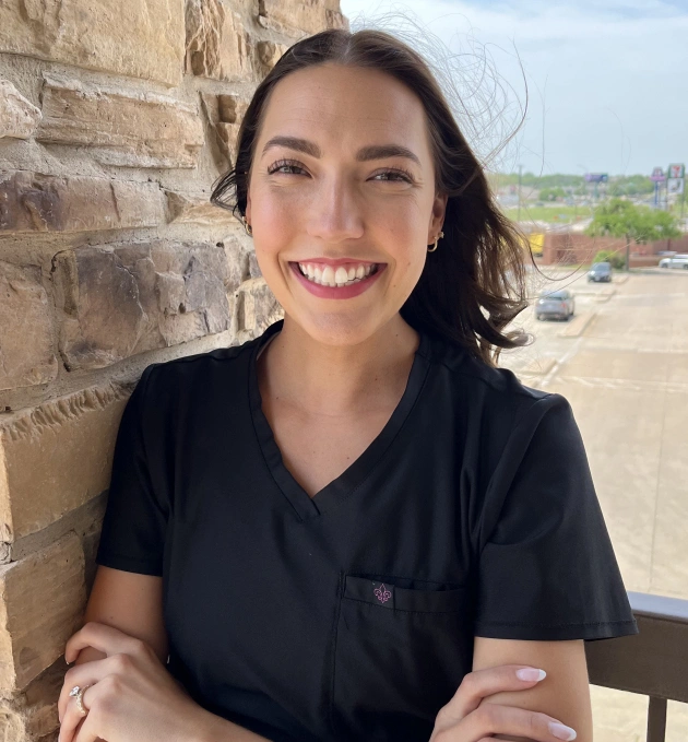 Woman in black scrubs smiling outdoors against brick wall at dental office.
