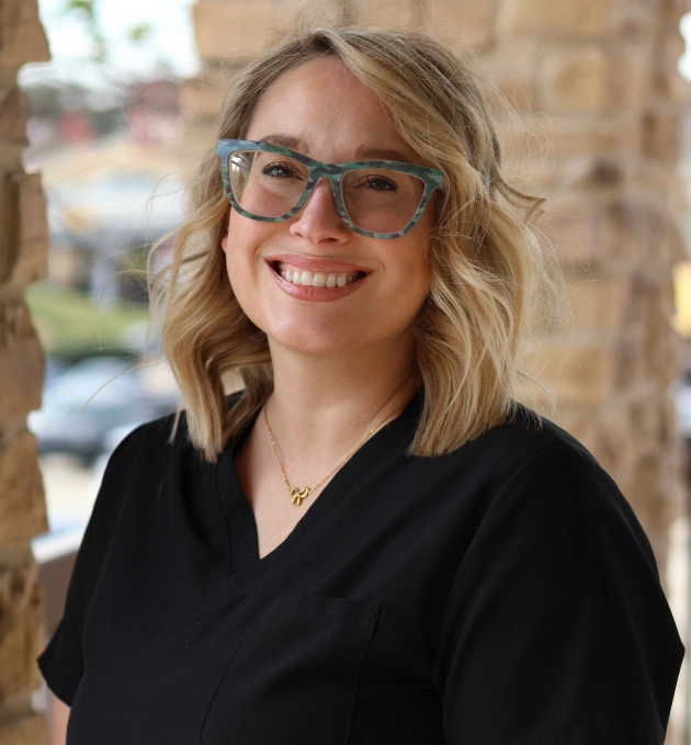 Professional headshot of smiling woman wearing glasses and black shirt in dental office setting.